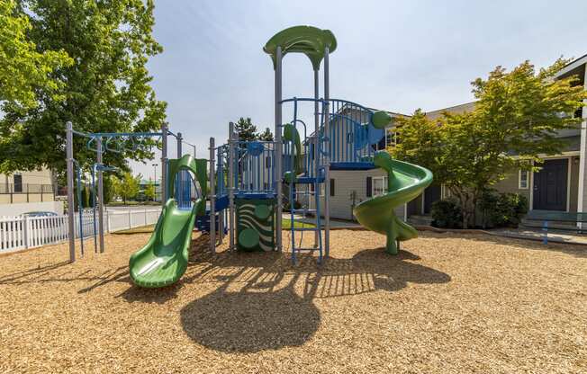 Green and blue playground surfaced with wood chips and enclosed by a white picket fence and green trees at Arterra Apartments, Kent, WA