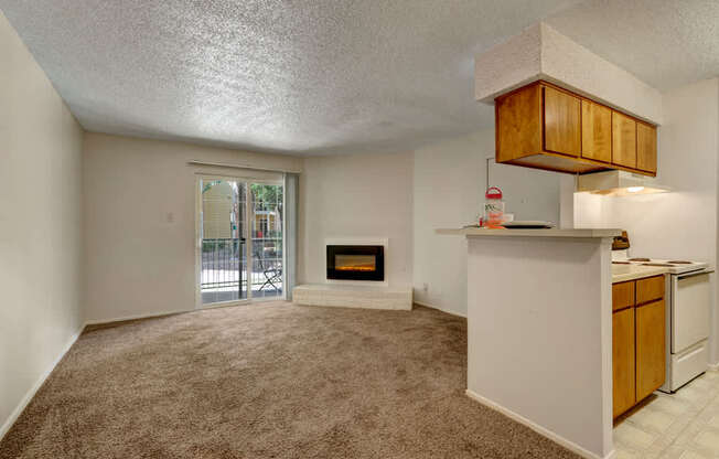 A spacious, empty living room here at The Bradford with plush beige carpet, neutral walls, and a cozy corner fireplace adding warmth. A sliding glass door brings in natural light and opens to an outdoor patio, while the open layout offers a clear view of the kitchen with warm wood cabinets and white appliances.
