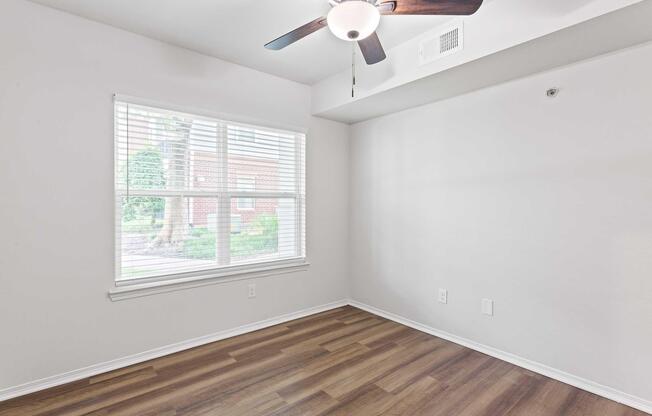 Bright, sparsely furnished room with light-colored walls and a ceiling fan. There's a window with blinds letting in natural light, revealing greenery outside. The flooring is wooden, and the space is ready for additional furniture or decoration.
