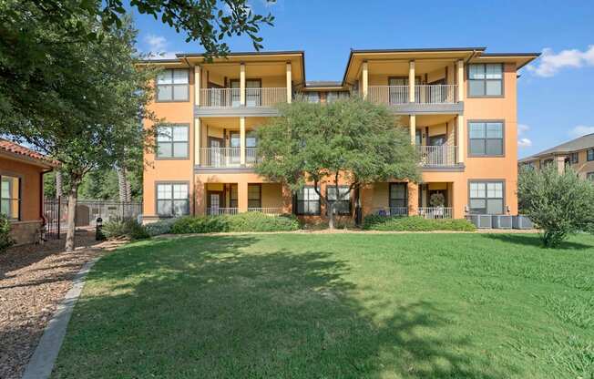 A well-maintained apartment building exterior here at Tuscany at Faudree featuring a three-story design with covered balconies, warm stucco tones, and large windows. Manicured green lawn, mature shade trees, and neatly landscaped walkways create a peaceful residential setting under a bright blue sky.