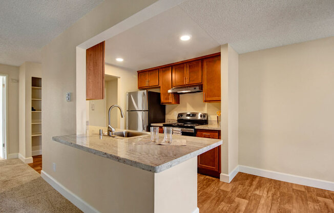 A bright open kitchen here at Adagio featuring warm wood cabinetry, light stone countertops, and a peninsula with a built-in sink and modern faucet, complemented by stainless steel appliances and a range hood, while wood-style flooring, recessed lighting, and an open connection to the living area create a functional, inviting space for cooking and entertaining.