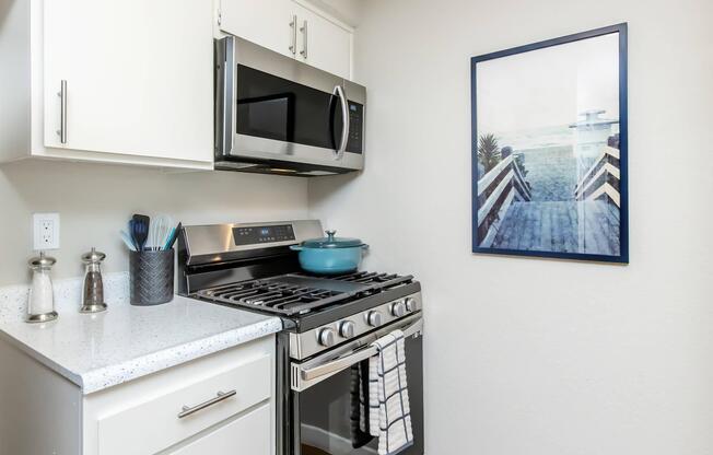 A modern kitchen featuring white cabinetry, a stainless steel microwave, a gas stove with a silver oven, and a blue cooking pot. The countertop is covered with a light speckled surface. A framed artwork of a beach scene hangs on the wall, adding a coastal touch to the space.