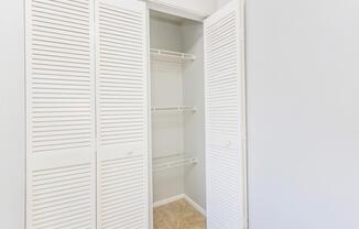 An empty closet with white louvered doors partially open. Inside, there are several wire shelves against a pale gray wall. The floor is tiled in a light beige color. The overall space appears bright and uncluttered, ideal for storage.