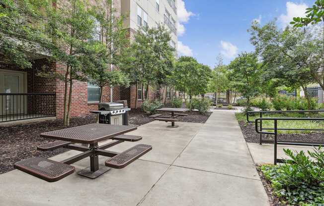 A concrete patio with a picnic table and benches is surrounded by trees and a brick building.