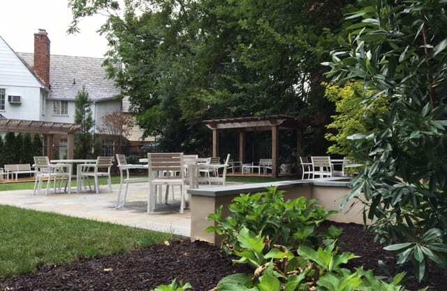 A patio with chairs and a table is surrounded by green plants.