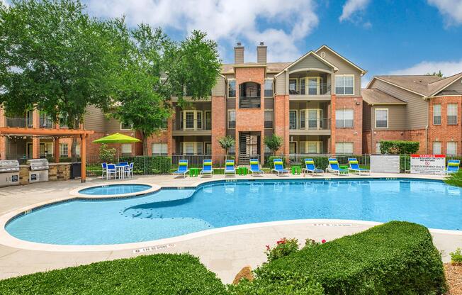 A swimming pool area surrounded by greenery, featuring lounge chairs and umbrellas. In the background, there are residential buildings with balconies and large windows. The scene is bright and sunny, with a clear blue sky. Outdoor barbecue grills are also visible near the pool.