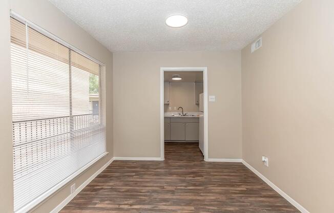 A well-lit, empty room featuring light-colored walls, a textured ceiling, and wooden flooring. A large window with blinds allows natural light in, and a doorway leads to a bathroom with a double sink visible in the background.