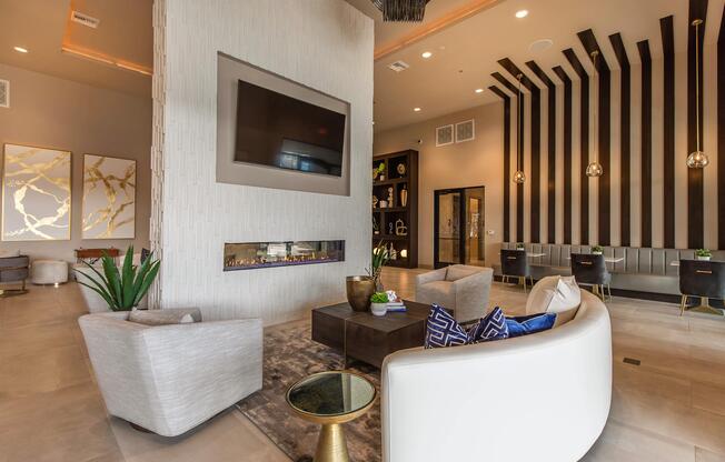Modern interior of a stylish living space featuring a sleek design. There is a cozy seating area with white armchairs and a round coffee table. A wall-mounted television is above a contemporary fireplace, accompanied by decorative artwork. The space has elegant lighting and wood accents.