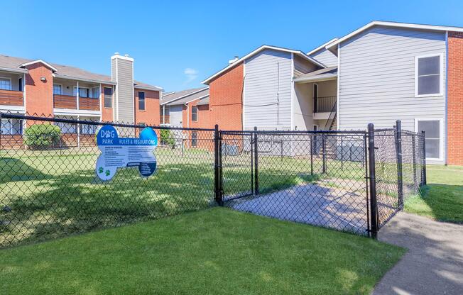 Fenced area labeled "Dog Park" with signage outlining rules and regulations, surrounded by grassy space and residential buildings in the background under a clear blue sky.
