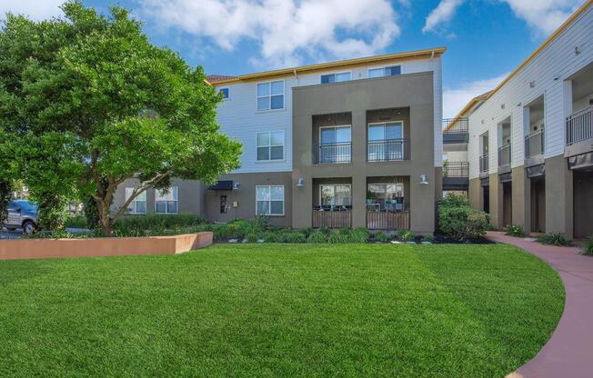 A modern apartment building with balconies, surrounded by lush green landscaping. In the foreground, a manicured lawn features low shrubs and trees. Partially cloudy blue sky overhead.
