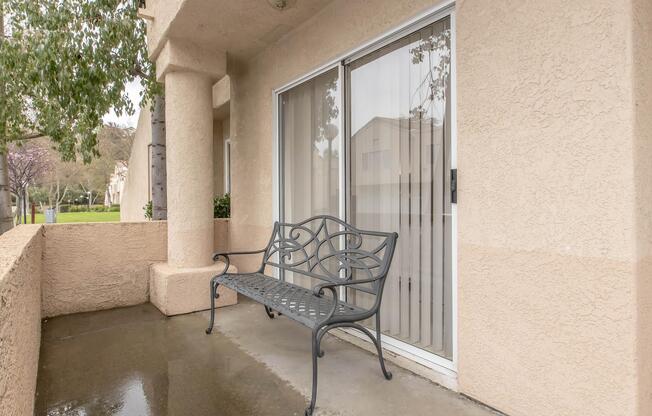 A small outdoor patio area featuring a metal bench near a sliding glass door. The patio is surrounded by textured beige walls, with a damp concrete floor and a view of greenery beyond.