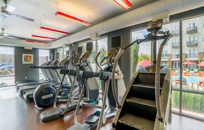 A modern fitness center here at BRIO at Firewheel with treadmills, ellipticals, and stair machines, large windows with pool views, and bright overhead lighting in an energizing workout space.