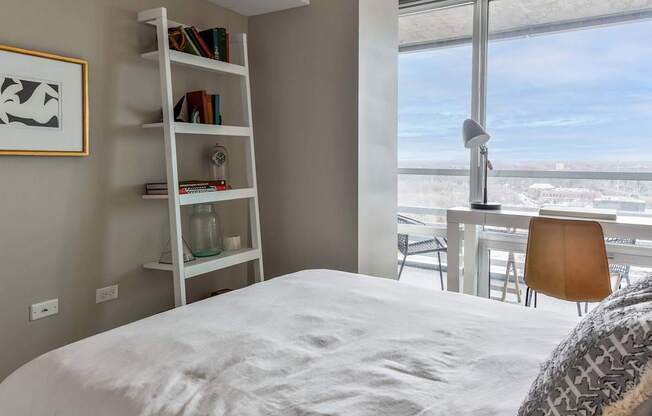 A white bedroom with a large bed, a desk with a chair, and a bookshelf with books and decorative items.