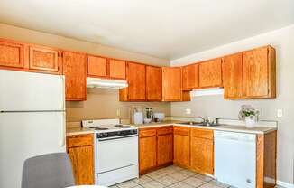 A simple kitchen with wooden cabinets, a white refrigerator, stove, microwave, and dishwasher on tiled flooring.