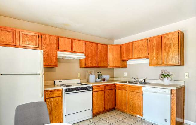 A simple kitchen with wooden cabinets, a white refrigerator, stove, microwave, and dishwasher on tiled flooring.