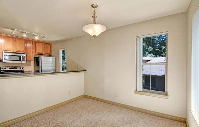 A neutral dining area here at Asbury Park featuring soft carpet flooring, a centered pendant light, and an open pass-through to the kitchen with a dark countertop, warm wood cabinetry, and stainless-steel appliances, while tall windows with vertical blinds bring in natural light and offer views of surrounding greenery.