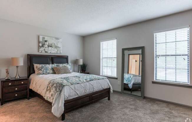 A bedroom with a large bed, a nightstand with a lamp, and a window with blinds.