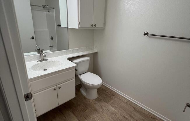 A clean, well-lit bathroom featuring a white countertop with a sink, a modern toilet, and a shower section. The walls are painted a light color, and there are white cabinets above the sink, with tiled flooring. A towel rack is mounted on the wall, contributing to the minimalist design.