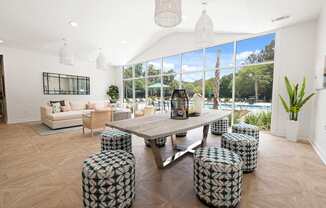 A modern clubhouse with a large table and ottomans Outside view of pool and clubhouse with pool furniture at The Palms at Chatham apartments in Savannah GA.