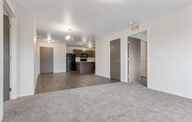 A spacious living room with a grey carpet and a kitchen area in the background.
