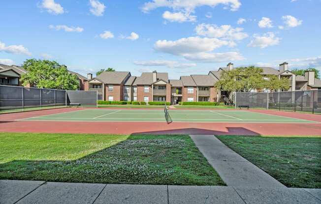 A tennis court is surrounded by a green lawn and apartment buildings.