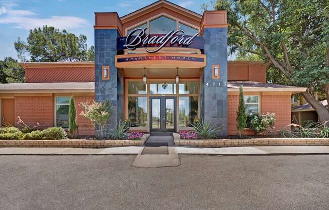 A welcoming clubhouse entrance here at The Bradford with a modern brick façade, tall glass doors, and clear property signage creating a polished first impression. A landscaped entry with greenery and flowers adds curb appeal and highlights the community’s inviting atmosphere.