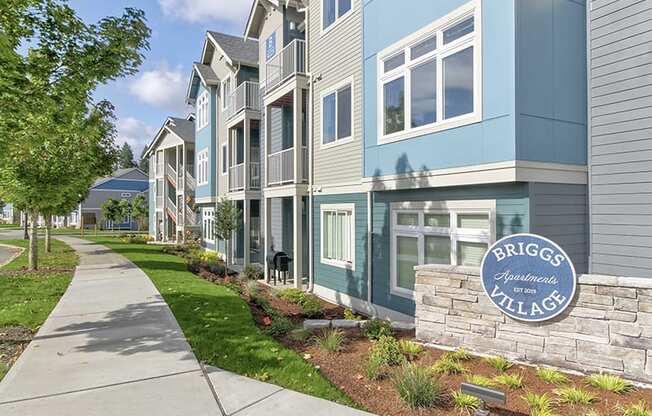 Apartments for Rent in Olympia - Briggs Village - Exterior Apartments Building with Grey and Blue Siding, Lush Greenery, and Sidewalk