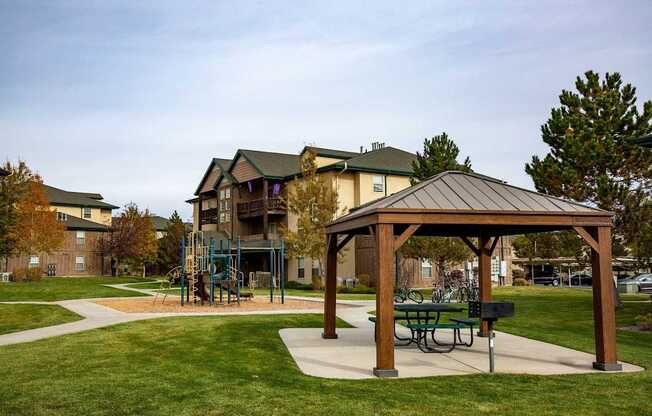 A wooden pavilion with a picnic table sits in a grassy area in front of apartment buildings.