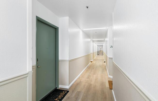 A long, well-lit hallway with white walls and a light wooden floor. Two doors are visible, one green and one white, with a welcome mat saying "hello" in front of the green door. The hallway has a clean, modern aesthetic with minimal decor.