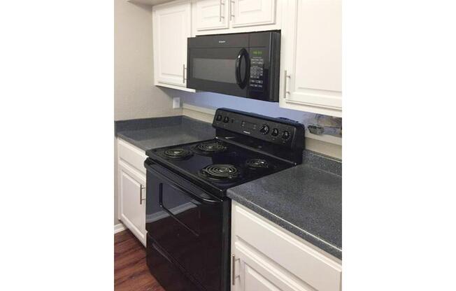 A modern kitchen featuring a black microwave mounted above a black electric stove with four burners. The countertops are a dark gray, and the cabinets above are white, creating a clean and sleek look. The flooring is a wooden design, adding warmth to the space.