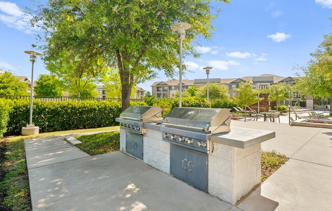 two stainless steel barbecue grills on a sidewalk in a park with trees