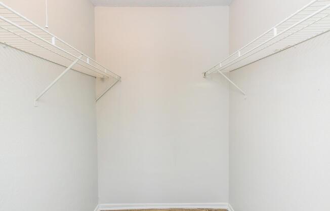 Empty closet with white walls and two wire shelves mounted on opposite sides. The floor is visible, showing a light-colored wood finish. The space is uncluttered, suggesting a potential for organization or storage.