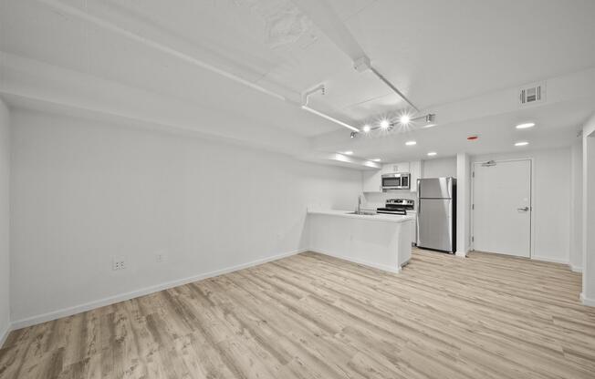 A spacious, modern studio apartment with light wood flooring. The walls are painted white, and there is a kitchenette equipped with stainless steel appliances. Recessed lighting illuminates the room, and the entrance door is visible in the background. The overall ambiance is bright and minimalistic.