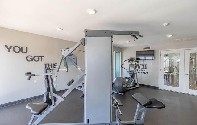 A modern gym interior featuring workout equipment, including a multi-station weight machine and cardiovascular machines, with motivational wording on the wall. Large windows provide natural light, enhancing the inviting atmosphere for fitness activities.