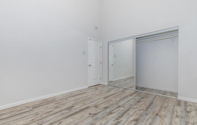 A spacious, empty room featuring light-colored walls and a high ceiling. The wooden floor has a warm tone, and there is a large mirror reflecting a closet area. A white door is visible on the left side of the image, enhancing the room's modern and minimalist aesthetic.