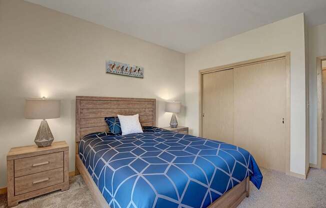A comfortable bedroom here at Vue at Pinnacle Park with soft carpet, a wood bed frame, blue patterned bedding, matching nightstands with lamps, and a wide sliding-door closet in a clean, cozy layout.