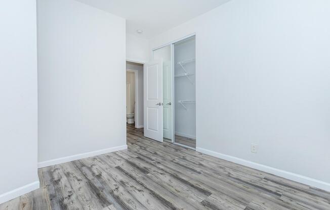 A clean, empty room featuring light-colored walls and a wooden floor. A partially open closet with sliding doors is visible on the left, and a doorway leads to another space in the background. The overall aesthetic is simple and minimalistic, providing a bright and spacious feel.