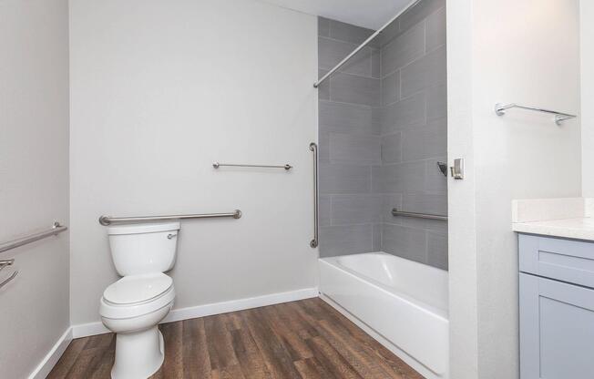 A modern, accessible bathroom featuring a toilet, a bathtub with a shower, and grab bars installed for safety. The walls are light-colored, and the flooring has a wood-like appearance, creating a clean and spacious atmosphere. A sink with a light blue cabinet is visible in the corner.