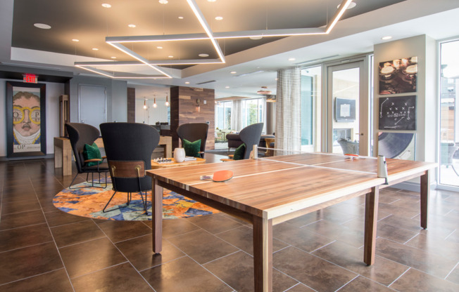 A lounge space with a ping pong table, four chairs around a coffee table, large windows, and colorful, angular modern art.