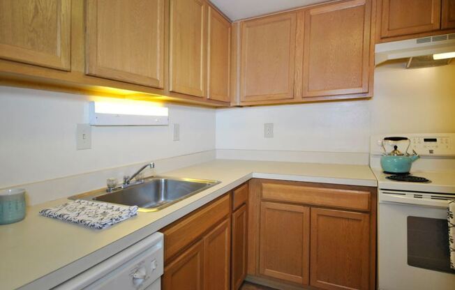 Photo of a kitchen, there are light wood cabinets below and above. The countertops ate white followed with white appliances. There is a sink and dishwasher to the left and a stove on the right. The walls are white and there are lights above the sink and oven.