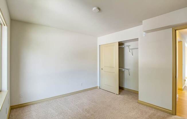 A comfortable bedroom here at Asbury Park featuring plush carpet flooring, soft neutral walls, and a large window that allows natural light to brighten the space, along with an open closet with shelving, a partially open interior door leading to the hallway, and ample wall space suitable for a bed, dresser, or additional storage.