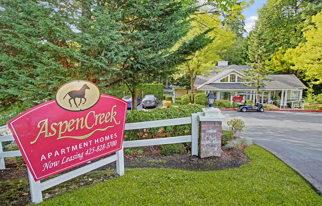 A landscaped community entrance here at Aspen Creek featuring a red Aspen Creek Apartment Homes sign set along a white fence, surrounded by green lawns, mature evergreen trees, and shrubs, with a paved drive leading toward the leasing office building visible in the background beneath a canopy of trees.
