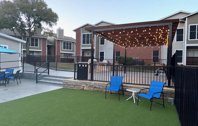 A patio area featuring two blue chairs and a small table on green turf, surrounded by black fencing. In the background, there are two brick buildings. A wooden pergola with string lights provides shade over the seating area, creating an inviting outdoor space for relaxation or gatherings.