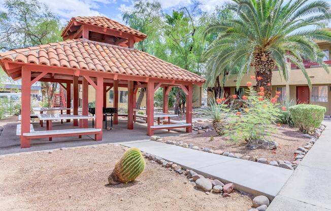 A shaded gazebo with a red tiled roof, surrounded by landscaped shrubs, palm trees, and gravel pathways. The area features picnic tables beneath the gazebo, offering a place for outdoor gatherings. A small cactus is visible in the foreground, adding to the desert ambiance.