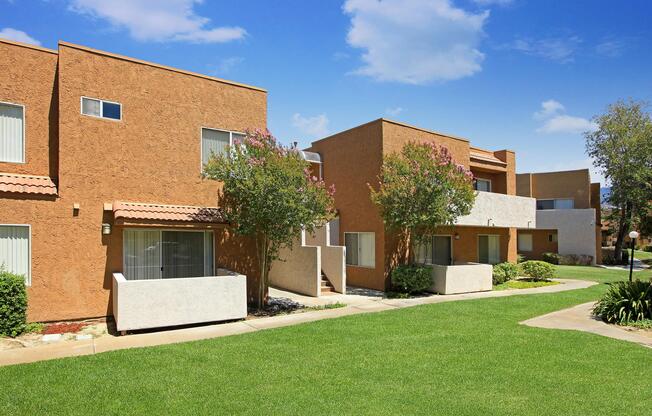 A landscaped residential area featuring two-story buildings with brown and beige exteriors. Each building is surrounded by green grass and small trees, with clear blue skies in the background. The scene conveys a peaceful, suburban atmosphere.