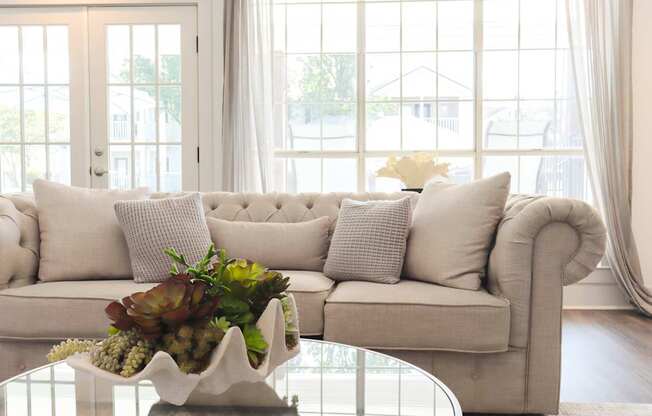 A white couch with a flower arrangement on a glass table in front of a window.