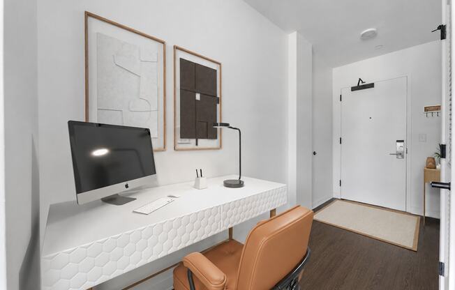 A white desk with a computer monitor, keyboard, and mouse is in a room with a brown chair and two framed pieces of artwork on the wall.