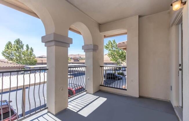 A view of a balcony featuring two archways, with a railing and shadow patterns on the floor. In the background, multiple cars are parked, and there are trees and a hillside visible under a clear blue sky.