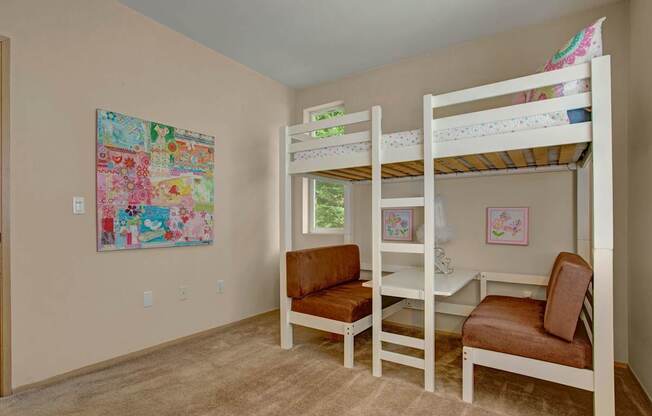 A playful bedroom here at Aspen Creek featuring soft neutral walls, plush carpet flooring, and a white loft bed with built-in ladder positioned over a small desk and seating area, accented by colorful wall art, framed prints, and a window that brings in natural light, creating a functional and cheerful space ideal for a child’s room or flexible use.