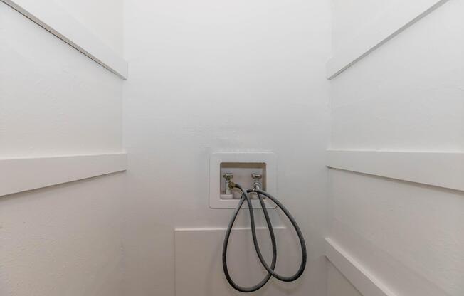 A narrow, white-walled space featuring a utility box with two hoses connected to it. The walls are flat and smooth, providing a clean, minimalistic look. The area appears empty and well-lit, emphasizing the utility connections.